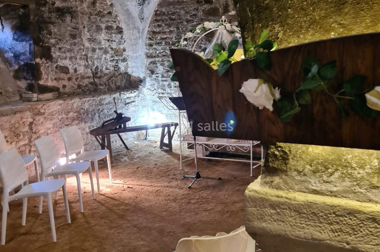 Salle voûtée en pierre avec des chaises blanches disposées pour un événement, décorée de fleurs et éclairée doucement.