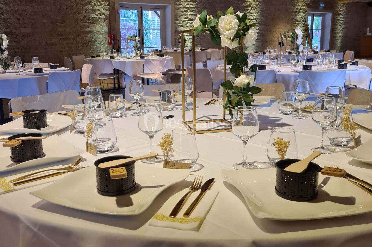 Salle de réception avec tables rondes dressées, nappes blanches, décorations florales et lumière naturelle.