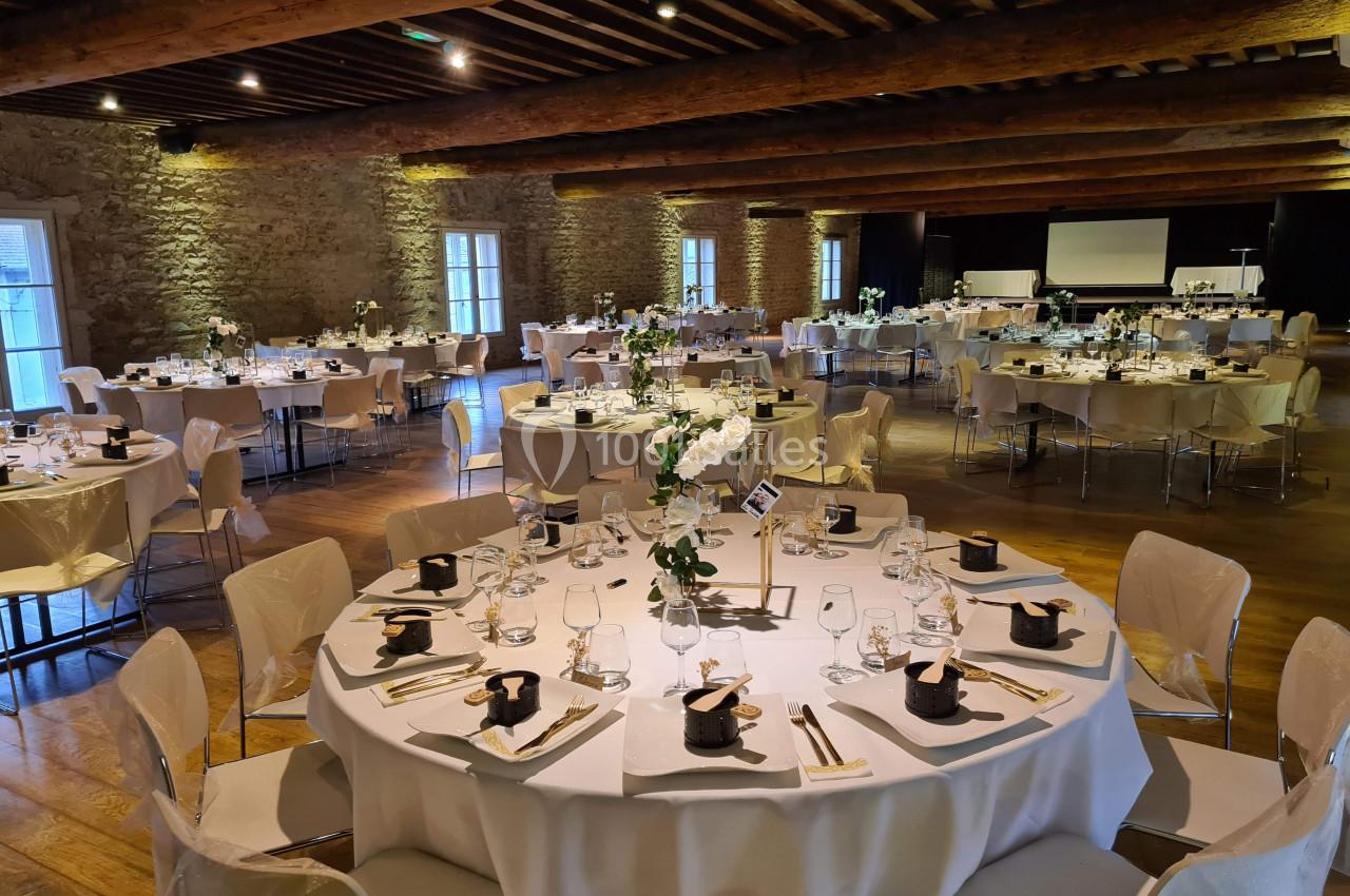 Salle de réception avec tables rondes dressées, nappes blanches, décorations florales et éclairage chaleureux.