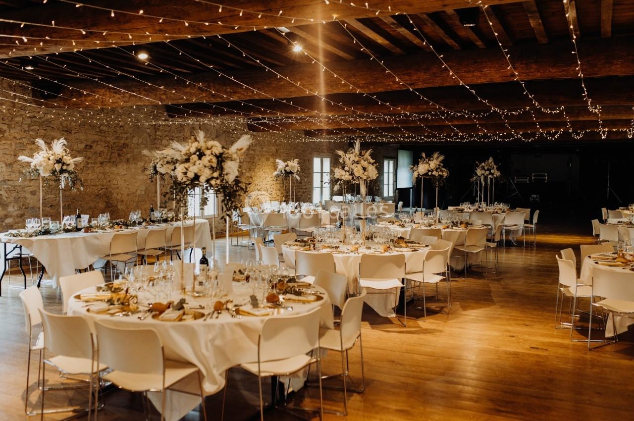 Salle de réception décorée avec des tables rondes, nappes blanches, centres de table fleuris et guirlandes lumineuses…