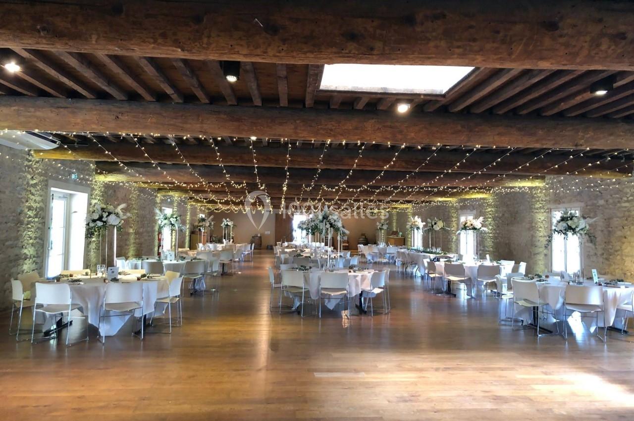 Salle de réception spacieuse avec tables décorées, poutres apparentes et guirlandes lumineuses suspendues.
