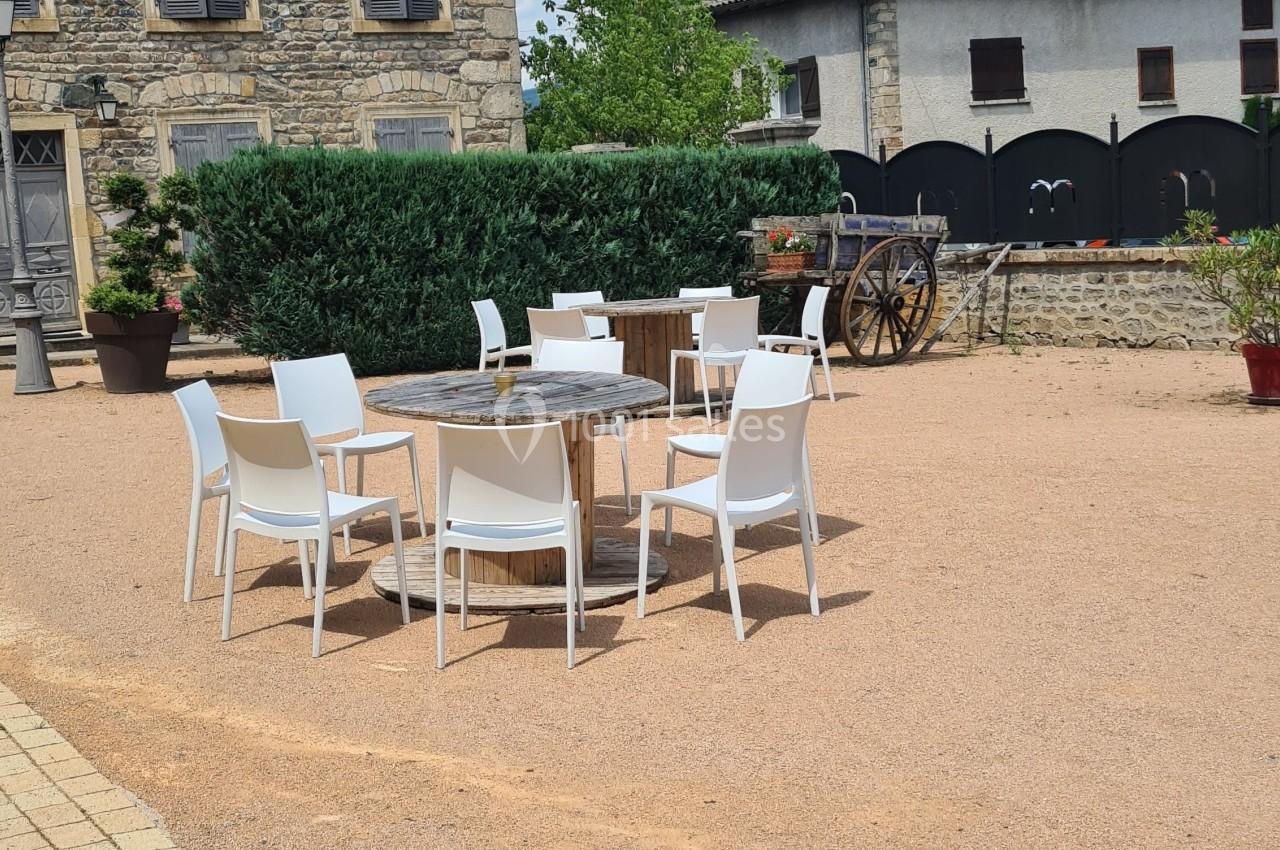 Chaises blanches et tables en bois disposées sur une place en gravier devant des bâtiments en pierre.