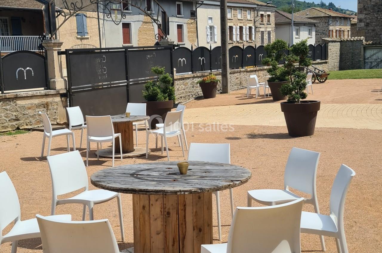 Chaises blanches disposées autour d'une table en bois sur une place ensoleillée avec des bâtiments en arrière-plan.