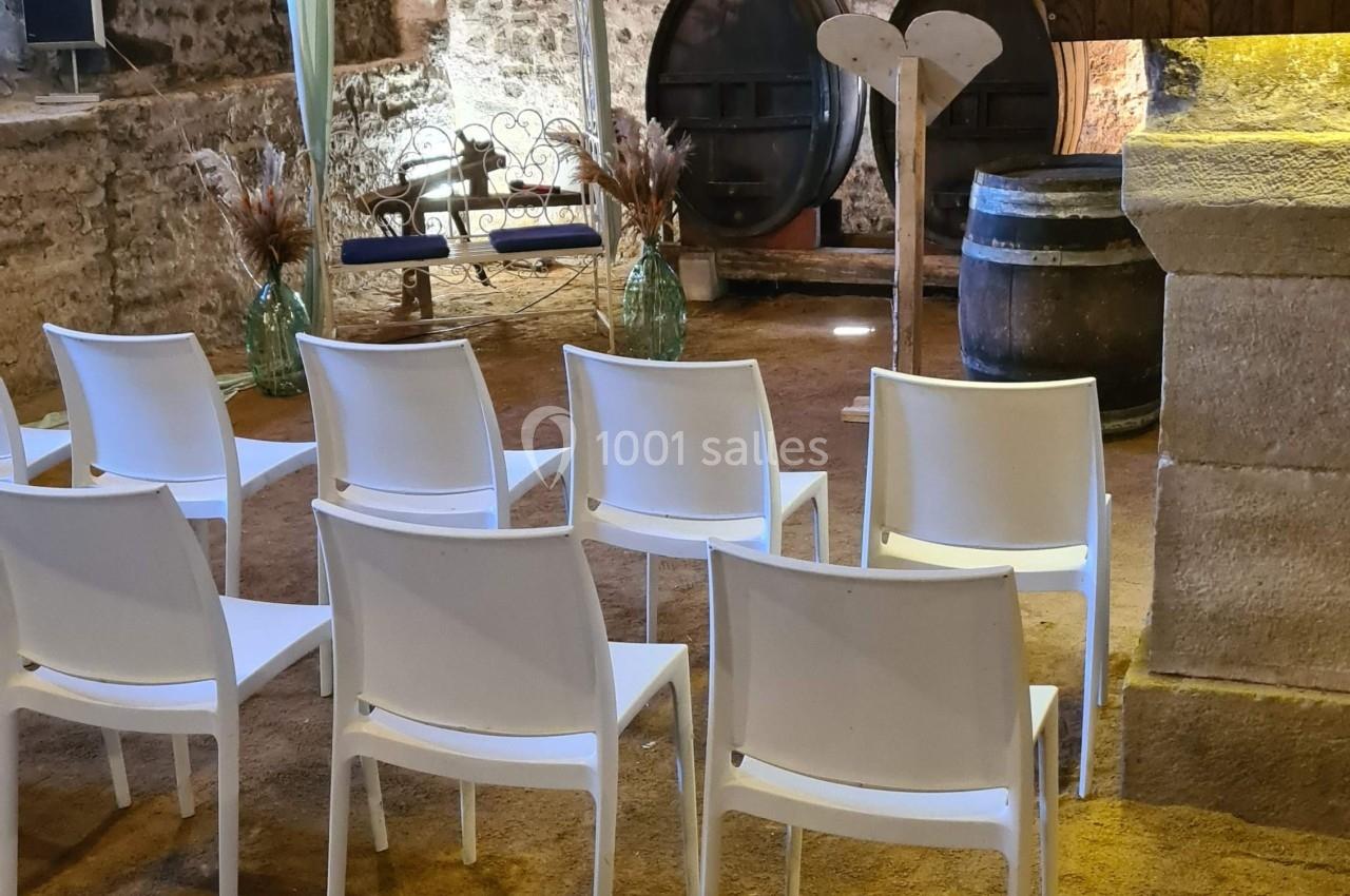 Salle voûtée en pierre aménagée pour une cérémonie, avec chaises blanches alignées et une arche décorative au fond.