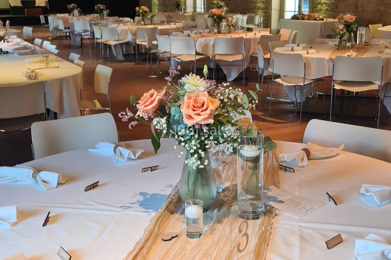 Salle de réception décorée avec des tables rondes, nappes blanches, centres de table fleuris et lumière naturelle.