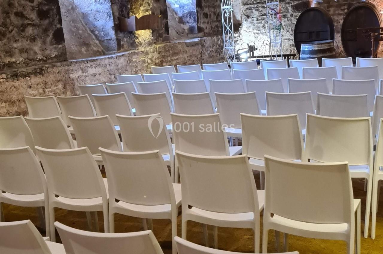Salle voûtée en pierre avec des rangées de chaises blanches disposées face à une arche décorative.