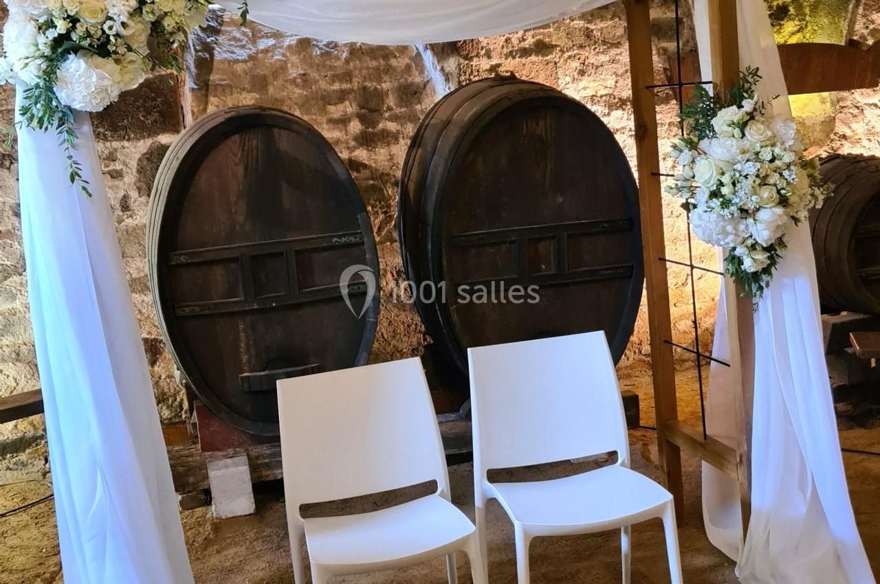 Arche décorée de fleurs blanches et de tissu, devant deux tonneaux en bois et deux chaises blanches, dans une cave voûtée.