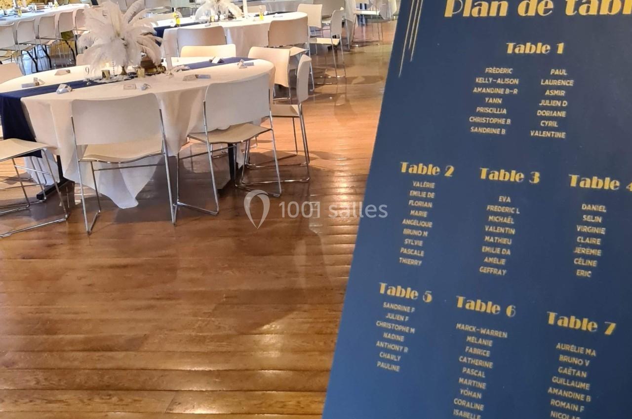 Plan de table sur un chevalet dans une salle de réception avec des tables rondes décorées et un parquet en bois.