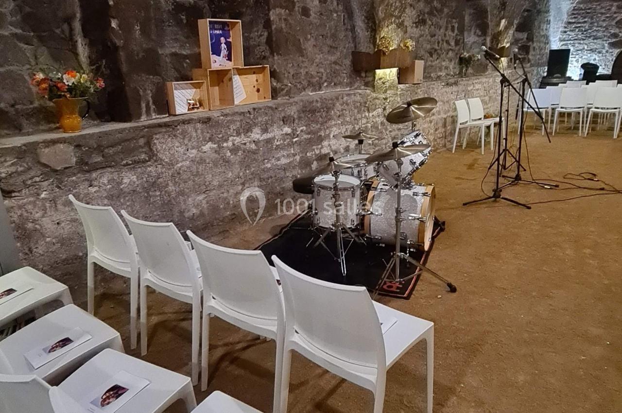 Salle voûtée en pierre avec chaises blanches alignées, une batterie, des micros et des décorations florales.