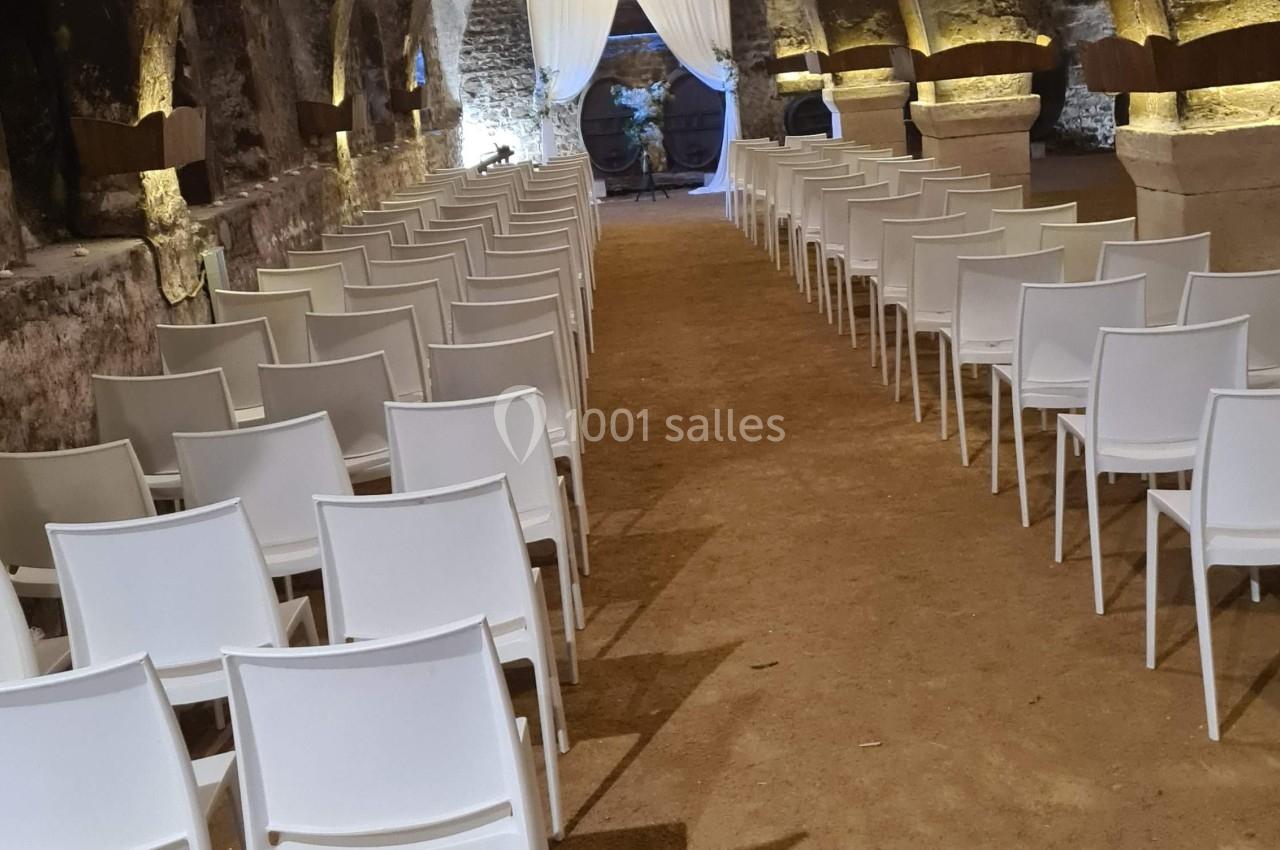 Rangées de chaises blanches alignées dans une salle voûtée en pierre avec un sol en terre battue.