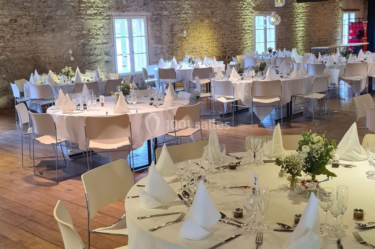 Salle de réception avec tables rondes dressées, nappes blanches, chaises modernes et murs en pierre apparente.