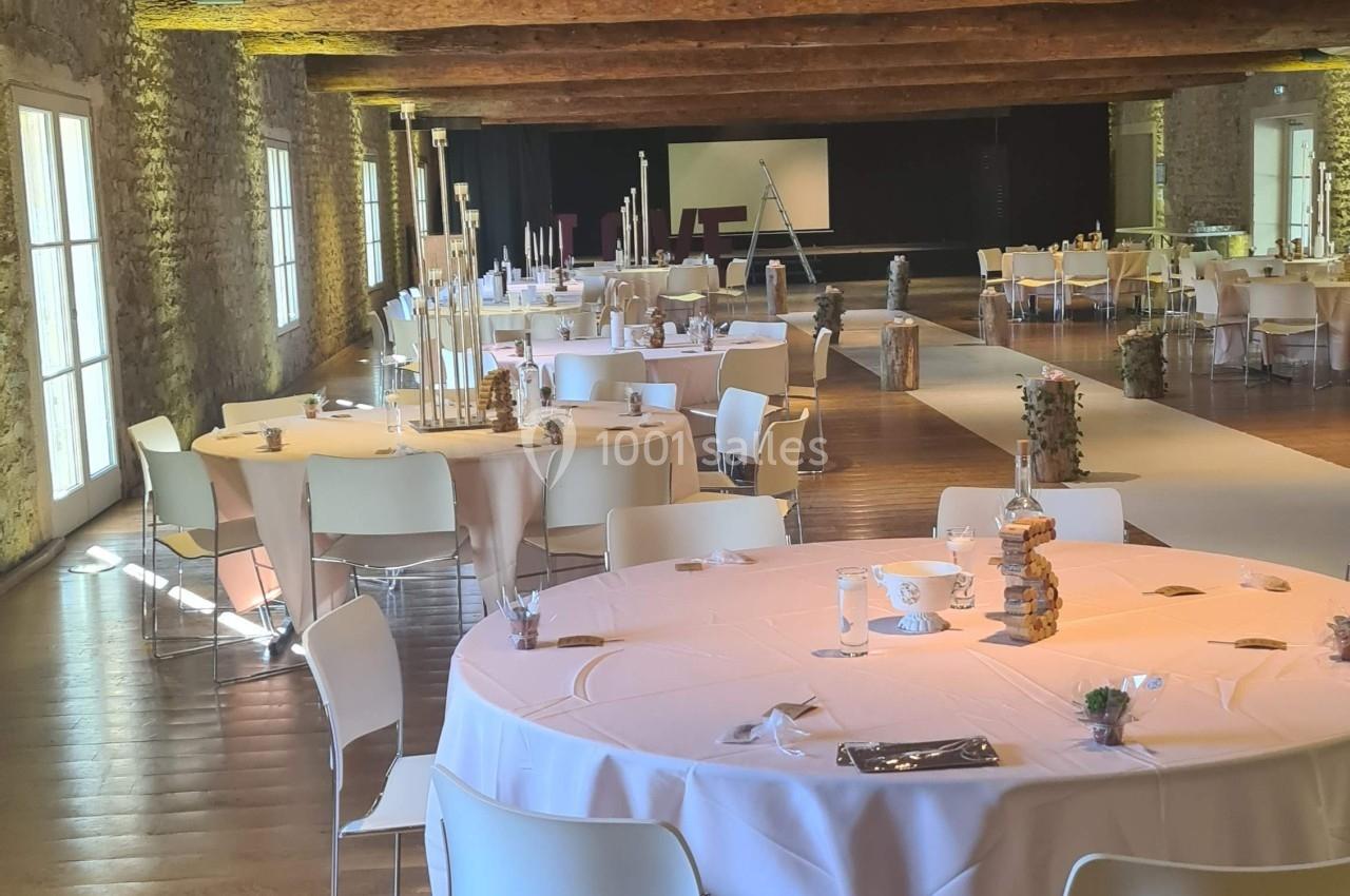 Salle de réception avec tables rondes décorées, chaises blanches et poutres apparentes au plafond.