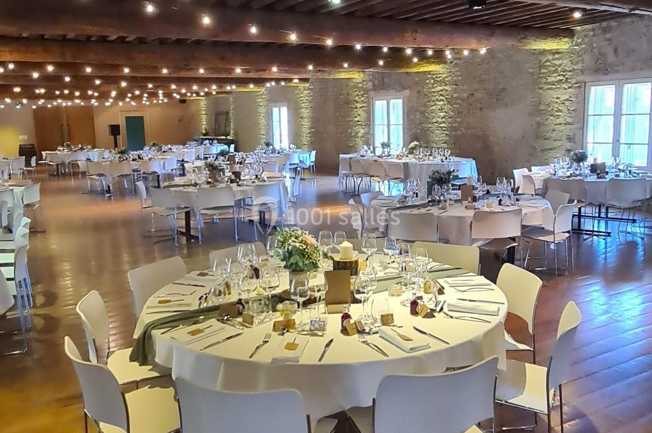 Salle de réception décorée avec des tables rondes dressées, chaises blanches et éclairage chaleureux sous un plafond en bois.