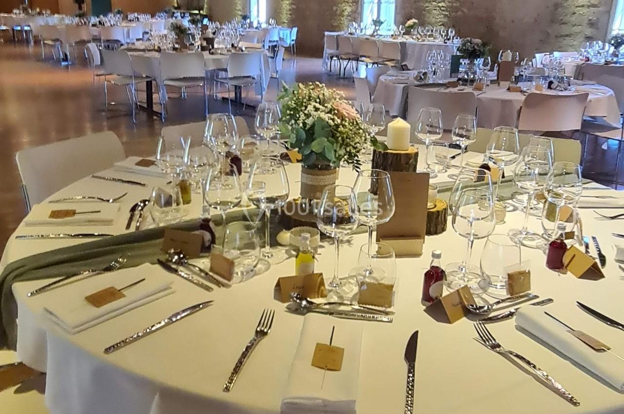 Salle de réception avec tables rondes dressées, décorées de fleurs, bougies et couverts élégants sous un plafond en bois.