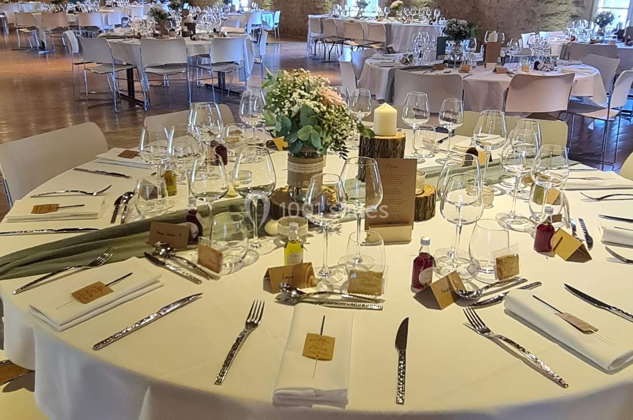 Salle de réception avec tables rondes dressées, nappes blanches, décorations florales et éclairage chaleureux.