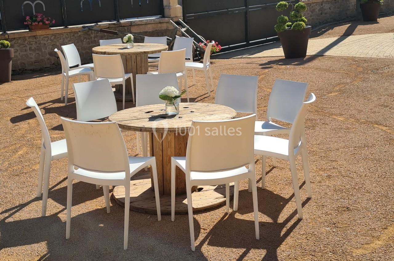 Chaises blanches disposées autour de tables en bois sur une terrasse extérieure en gravier, avec des bâtiments en arrière…