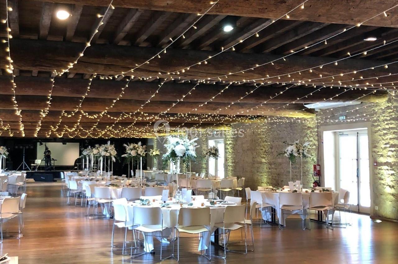 Salle de réception décorée avec des guirlandes lumineuses, tables dressées et poutres apparentes en bois.