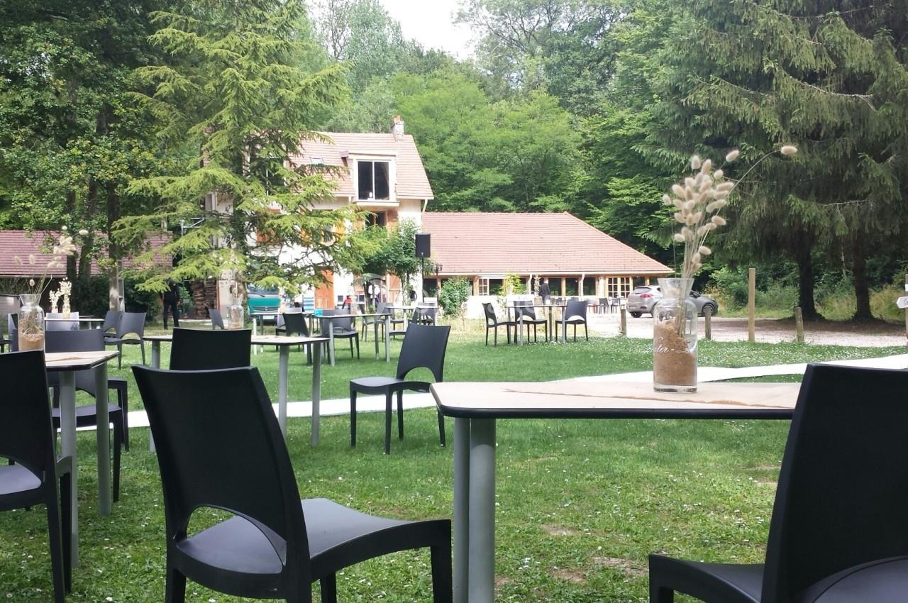 Terrasse extérieure avec tables et chaises noires, entourée de verdure et proche d'une maison au toit en tuiles.