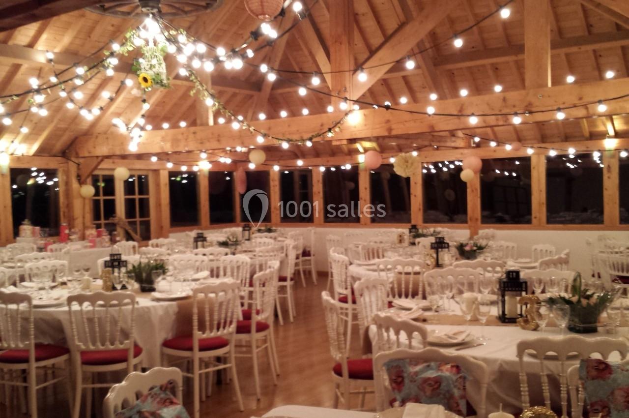 Salle de réception en bois décorée de guirlandes lumineuses, tables rondes dressées avec nappes blanches et chaises.