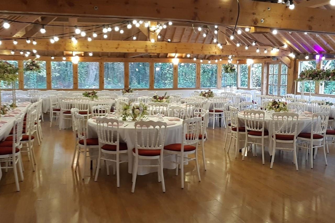 Salle de réception décorée avec des tables rondes, nappes blanches, chaises blanches et guirlandes lumineuses suspendues.