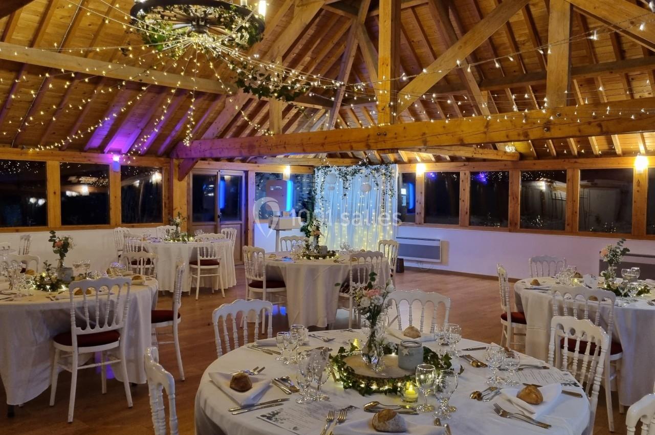 Salle de réception décorée avec des guirlandes lumineuses, des tables rondes dressées et un plafond en bois apparent.