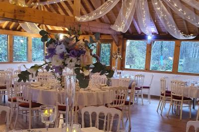 Salle de réception décorée avec des guirlandes lumineuses, des tables rondes dressées et un plafond en bois apparent.