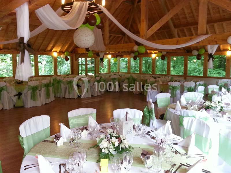 Salle de réception décorée avec des tables rondes, nappes blanches et vertes, et guirlandes suspendues au plafond en bois.