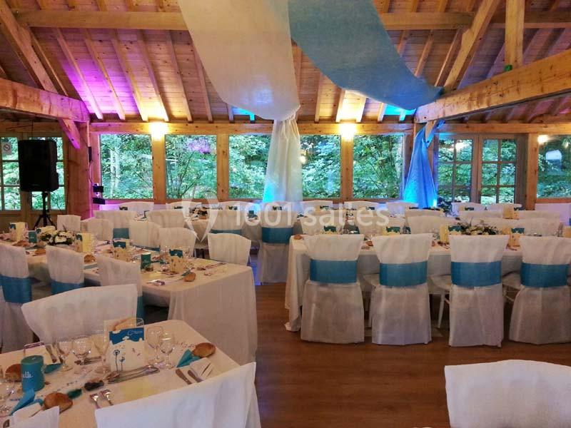 Salle de réception en bois décorée pour un événement, avec tables dressées et chaises ornées de rubans bleus.