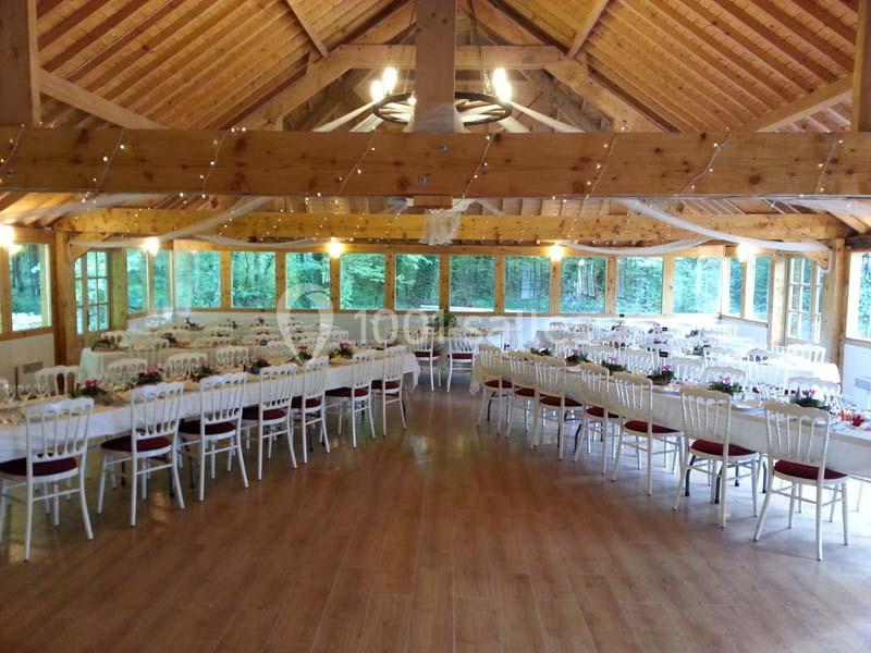 Salle de réception en bois avec tables décorées et chaises blanches, entourée de grandes fenêtres donnant sur la nature.