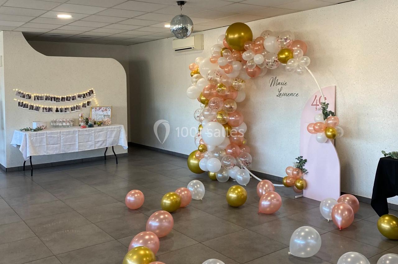 Décoration de salle avec arche de ballons roses, blancs et dorés, table décorative et guirlande de photos au mur.