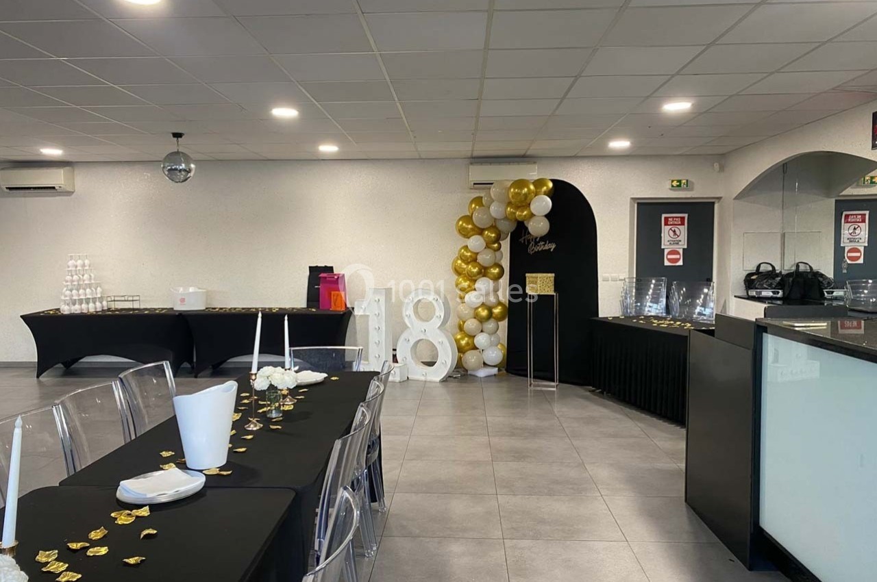 Salle décorée pour une fête avec tables noires, chaises transparentes, ballons dorés et blancs, et chiffre 18 en arrière…