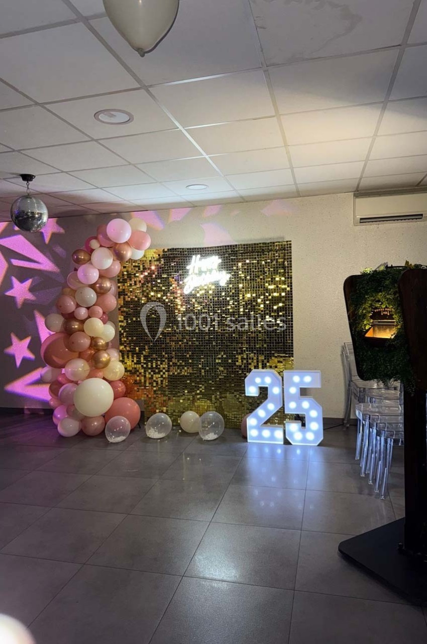 Décoration festive avec mur scintillant, ballons roses et blancs, chiffre lumineux ’25’ et éclairage tamisé.