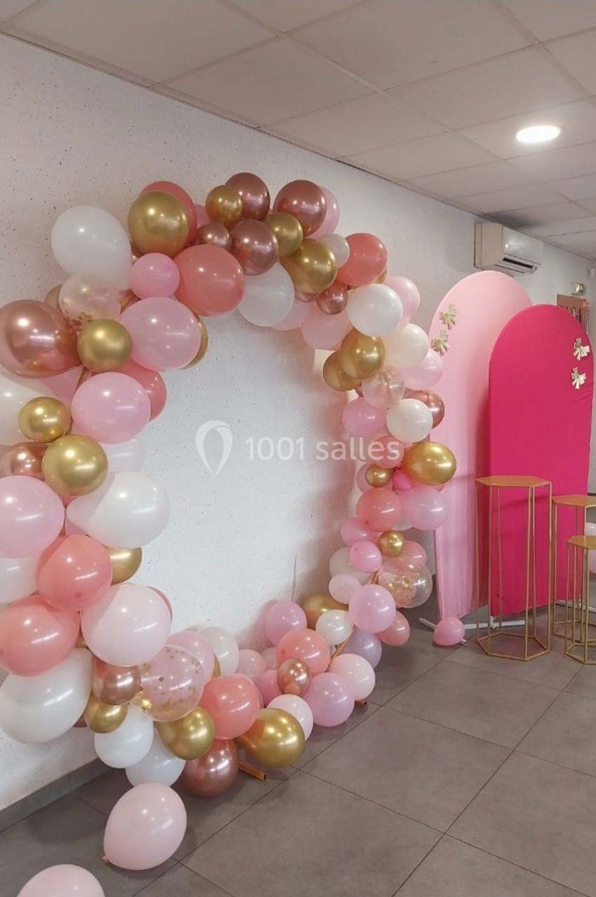 Arc de ballons roses, blancs et dorés décorant un espace intérieur avec un fond rose et des tabourets dorés.
