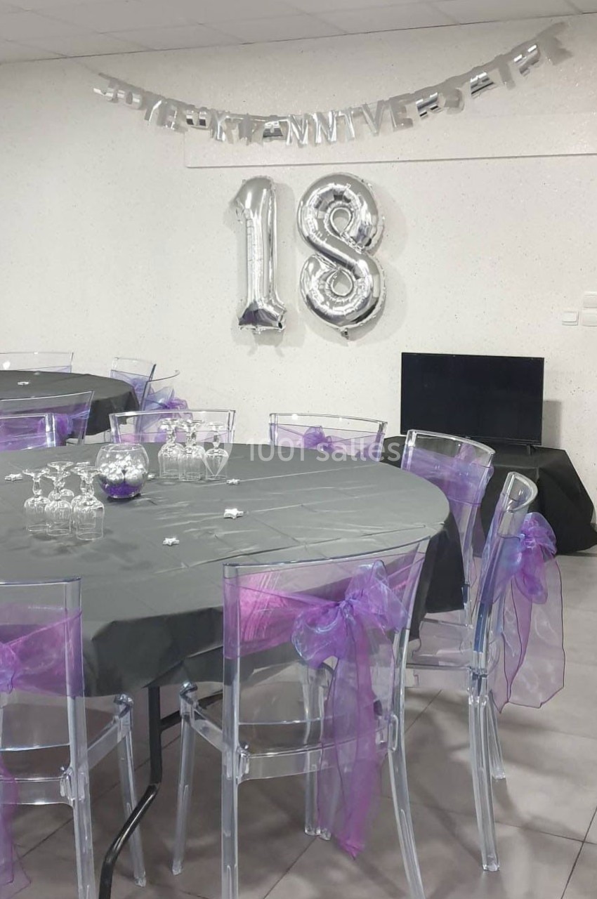 Salle décorée pour un 18e anniversaire avec des ballons argentés, nappes grises et chaises ornées de rubans violets.