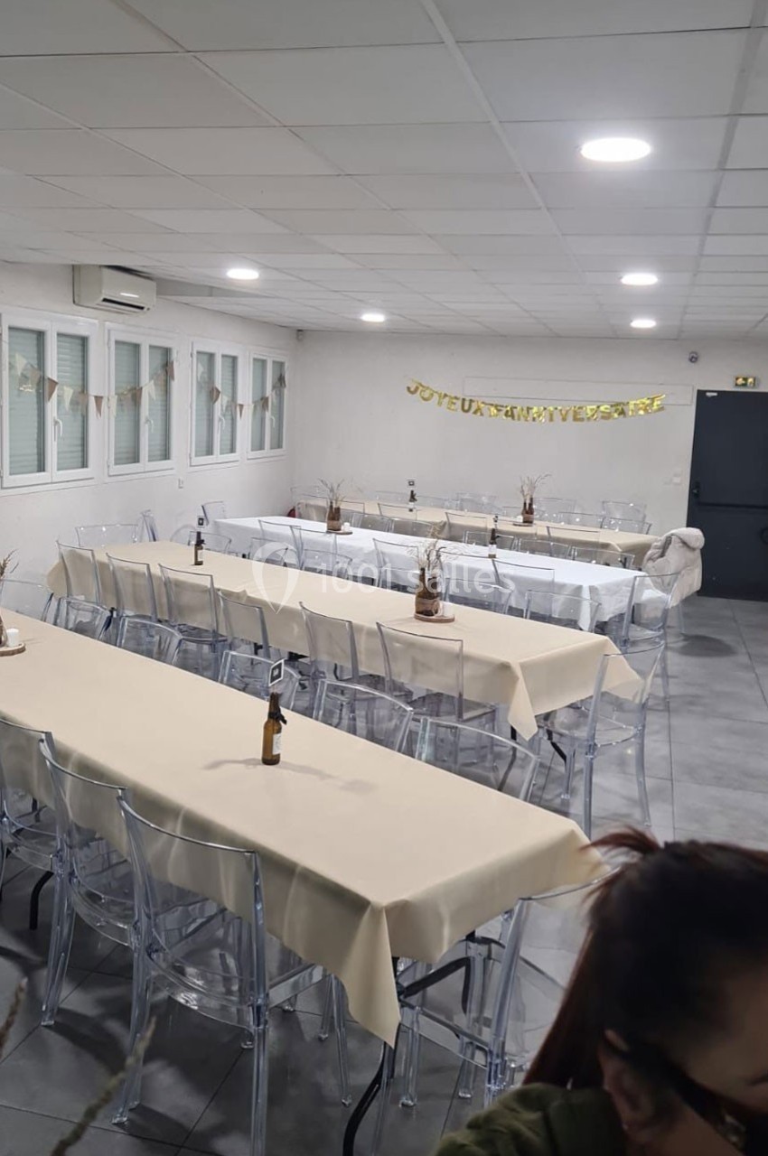 Salle décorée pour un anniversaire avec des tables alignées, nappes beiges, chaises transparentes et guirlande festive.
