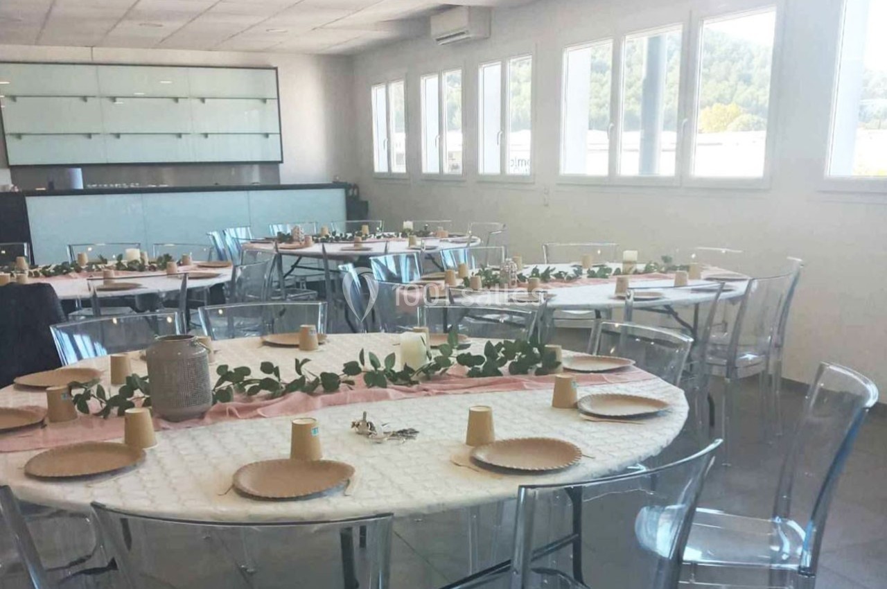 Salle lumineuse avec des tables rondes dressées pour un repas, décorées de nappes blanches et de chemins de table roses.