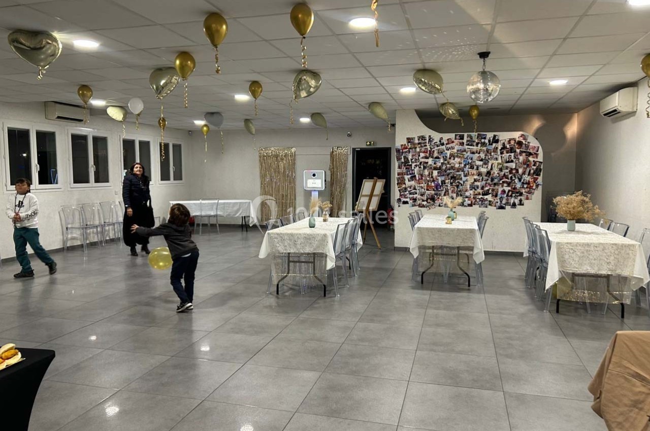 Salle décorée avec des ballons dorés, tables dressées et mur couvert de photos, avec des enfants jouant au sol.