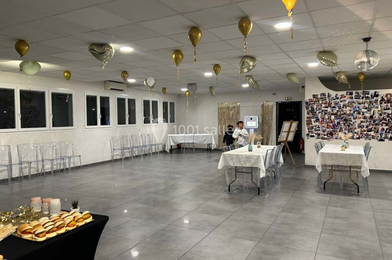 Salle décorée pour un événement avec ballons dorés au plafond, tables dressées et buffet en premier plan.