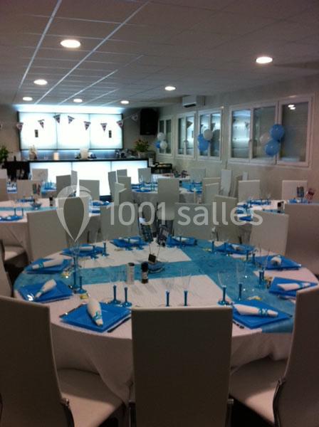Salle décorée pour un événement, avec des tables rondes dressées en bleu et blanc, et des ballons en arrière-plan.