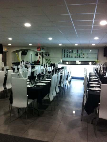 Salle de réception avec tables dressées, nappes noires, chaises blanches et un bar éclairé en arrière-plan.