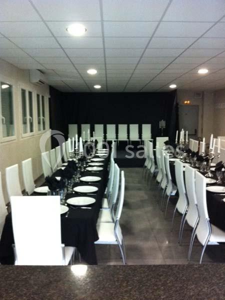 Salle de réception avec tables alignées, nappes noires, chaises blanches et chandeliers disposés pour un événement.