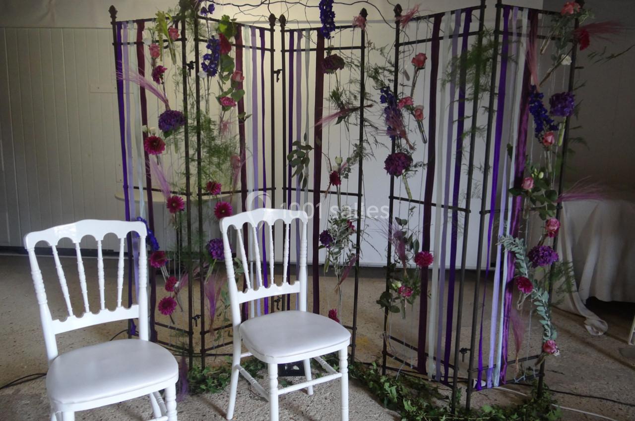 Deux chaises blanches devant une structure métallique décorée de fleurs, feuillages et rubans violets.
