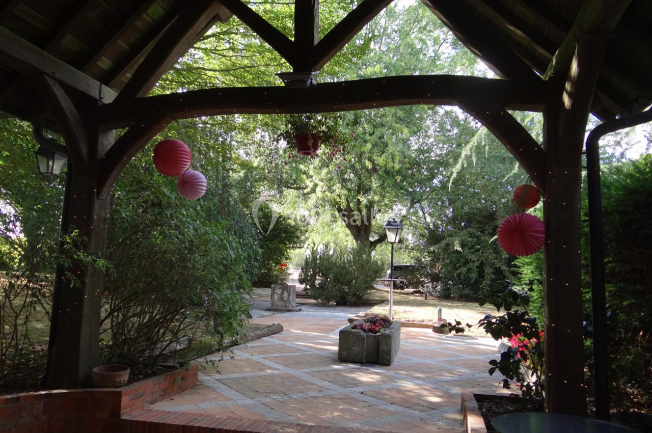 Vue d'une terrasse couverte en bois donnant sur un jardin arboré avec des lanternes suspendues et des parterres fleuris.