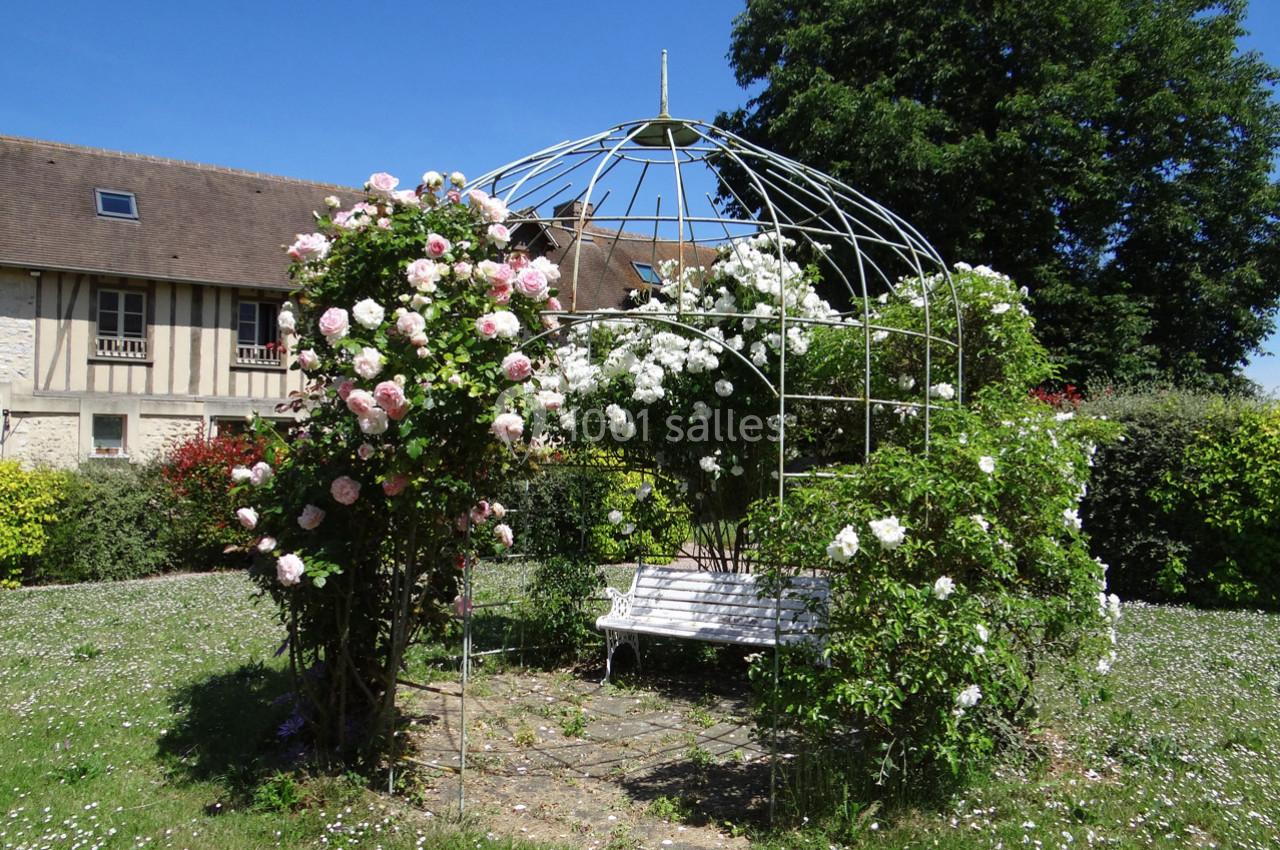 Tonnelle en métal recouverte de rosiers fleuris, avec un banc blanc, dans un jardin ensoleillé près d'une maison à…