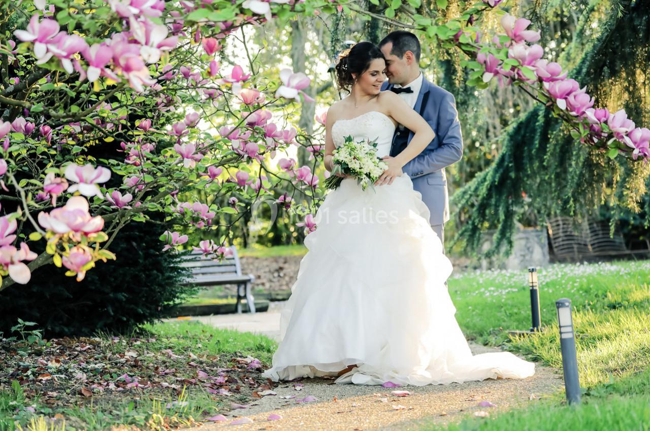 Un couple de mariés pose dans un jardin fleuri, entouré de magnolias roses et d'une lumière douce.