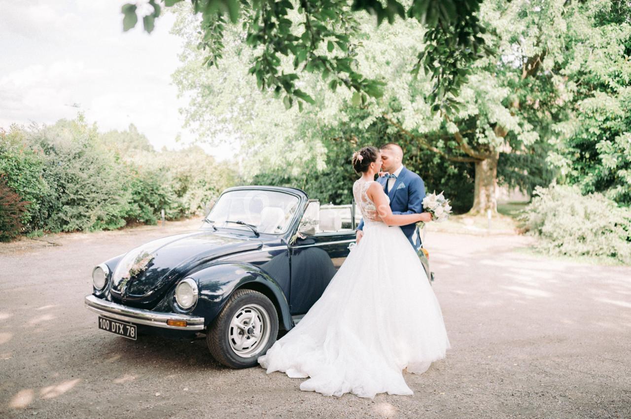 Un couple de mariés s'embrasse près d'une voiture décapotable noire dans un cadre verdoyant et ensoleillé.