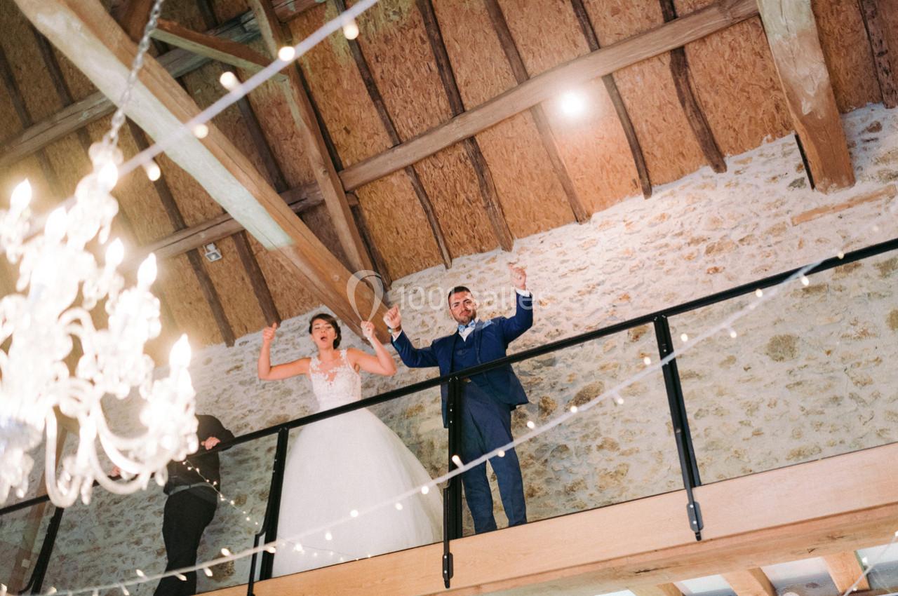 Un couple en tenue de mariage se tient sur une mezzanine en bois, sous un plafond avec poutres apparentes et guirlandes…