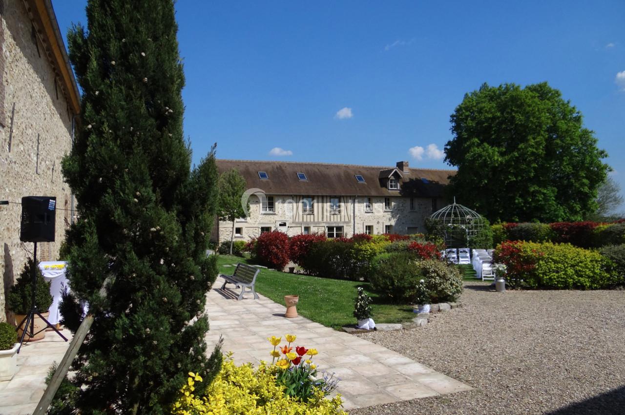 Cour d'une propriété en pierre avec jardin fleuri, allée gravillonnée, et tonnelle sous un ciel bleu.