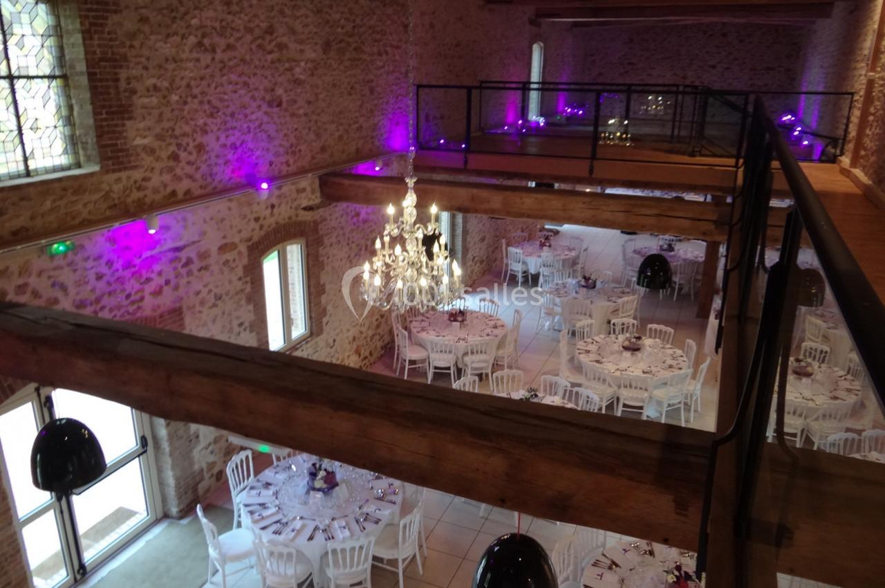 Salle de réception en pierre avec tables rondes dressées, lustre central et éclairage violet tamisé.