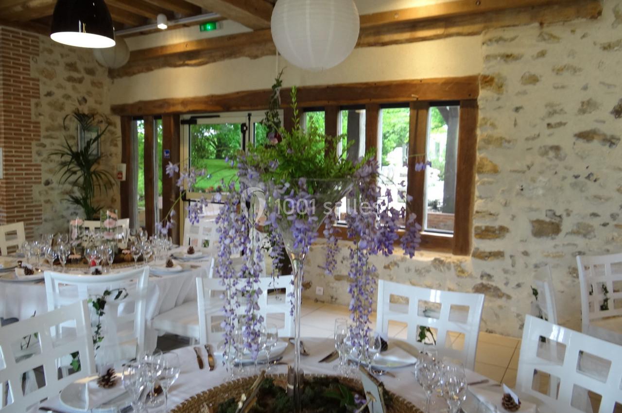 Salle de réception lumineuse avec tables dressées et centre de table floral composé de glycines violettes.